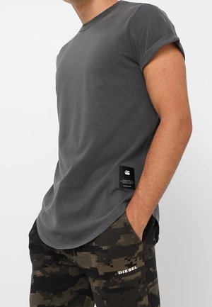 Short-sleeve, fitted gray t-shirt with a round neckline and rolled cuffs. Features a branded label near the hem. Worn with camo-patterned pants.