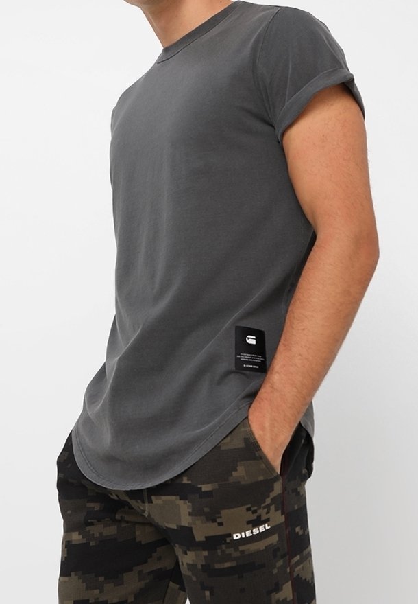 T-shirt gris à manches courtes, ajusté, avec un col rond et des revers. Présente une étiquette de marque près de l'ourlet. Porté avec un pantalon à motif camouflage.
