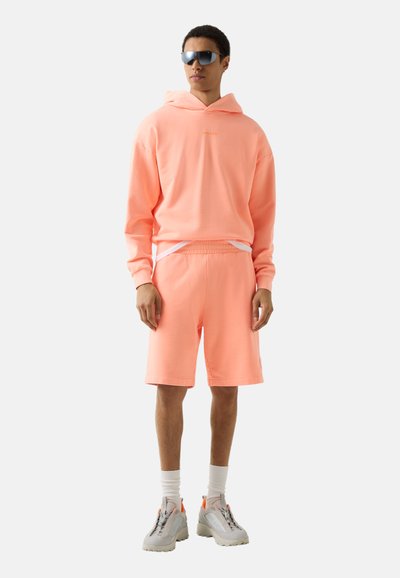 Ung person iført ferskenfarvet hættetrøje, matchende shorts, hvide sokker, chunky sneakers og sorte solbriller, stående mod en ensfarvet baggrund.