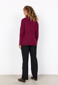 Burgunderfarbener ribbeliger Strickpullover mit Rundhalsausschnitt, langen Ärmeln und Seitenschlitzen, kombiniert mit schwarzen geraden Hosen und braunen Punkte-Ballerinas.
