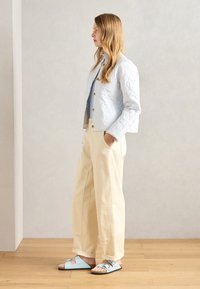 Svetlo modra prešita jakna z zlatimi gumbi, oblečena čez svetlo modro majico, v kombinaciji z bež širokimi hlačami in svetlo modrimi sandali.