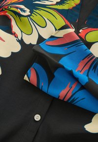 Chemise noire avec de grands motifs floraux colorés en bleu, rouge, vert et crème, montrant un bouton blanc et un détail en tissu plié.