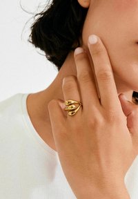 Kuldne sõrmus, millel on kolmelehelehele iseloomulik disain kõverate kujudega ja siledad, peegeldavad pinnad. Käsi on esitletud tekstuurse valge topi taustal.