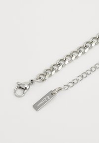 Cadena de metal plateado con un cierre de langosta, que presenta un diseño de eslabones planos y una etiqueta rectangular con el logo. Textura suave y acabado pulido.