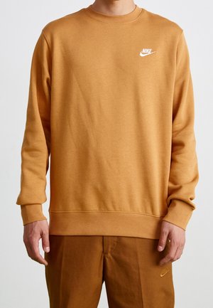 Osoba ubrana w brązową bluzę Nike i dopasowane spodnie, stojąca z ramionami swobodnie opuszczonymi wzdłuż ciała na jednolitym tle.