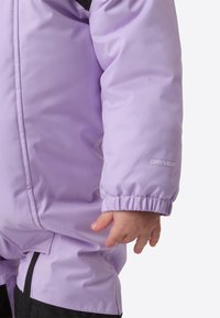 Fialová zimní bunda z voděodolného materiálu. Má elastické manžety a logo Dryvent, na detailním záběru ruka držící tkaninu.
