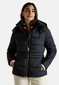 Žena v čiernej prešívanej bunde s červeným a bielym pruhovaným sveterom, s rukami v vreckách na pozadí bez vzoru.