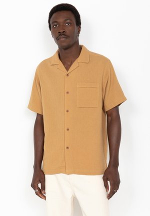 Mand iført en kortærmet, sennepsfarvet skjorte med knapper og brystlomme samt off-white bukser, stående foran en hvid baggrund.