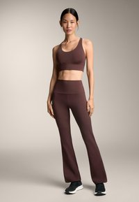 Brązowy stanik sportowy i spodnie z wysokim stanem, wykonane z gładkiego, elastycznego materiału. Modelka stoi w czarnych trampkach na neutralnym tle.