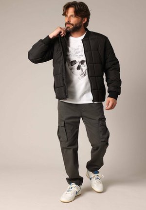 Veste noire en duvet avec un motif de carrés matelassés, t-shirt graphique blanc avec un crâne, pantalon cargo sombre et baskets blanches avec des accents bleus.