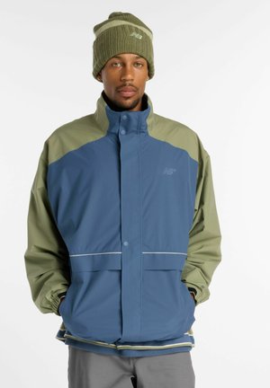 Mann trägt eine blau-olivgrüne Jacke und passende olivgrüne Strickmütze und steht mit den Händen in den Taschen vor weißem Hintergrund.