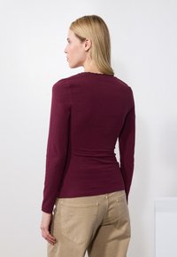 Burgundy long-sleeve top with a fitted design, round neckline, and gathering at shoulders; paired with light beige pants.