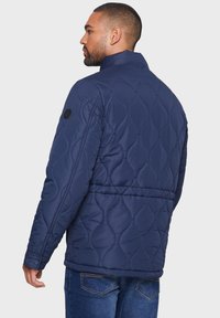 Marineblå quilted jakke med høj krave, der har et struktureret mønster og en sort detalje på skulderen. Parret med blå jeans.