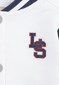 Fehér pulóver sötétkék kiegészítőkkel, kerek nyakkal és gombos záródással. Figyelemre méltó "LS" logó sötétkék és piros színben az elején.