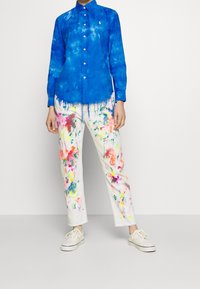 Person som bär en klarblå långärmad skjorta med knappar och vita byxor med färgglada färgsplatterdesigns, kombinerat med vita sneakers.