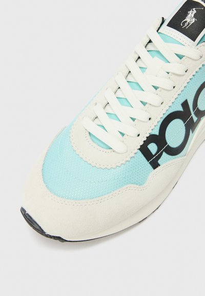 Adidași din țesătură albastru deschis cu accente din suede crem, având șireturi albe, un logo mare negru "POLO" și un design cu vârful rotunjit.