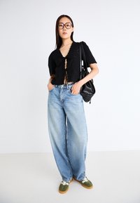Černý krajkový crop top s krátkými nabíranými rukávy a zavazováním vpředu, spárovaný se světle modrými širokými džíny a olivovými teniskami. Černá kabelka přes rameno.