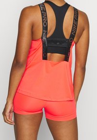 Haut de sport sans manches de couleur corail vif avec des accents noirs. Il présente un dos ajouré et des bretelles texturées avec branding. Porté avec un short assorti.