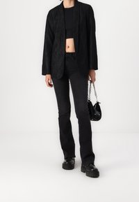 Personne portant un blazer en dentelle noire sur un top noir court, un jean noir, des chaussures à plateforme noires, tenant un petit sac à main noir avec une bandoulière en chaîne.