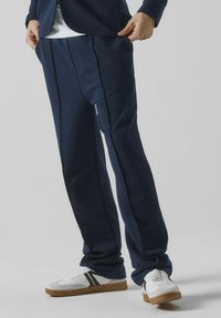 Navyblå joggerbyxor i mjukt material, med veck och sid sömmar, tillsammans med retroinspirerade vita sneakers med svarta accenter.