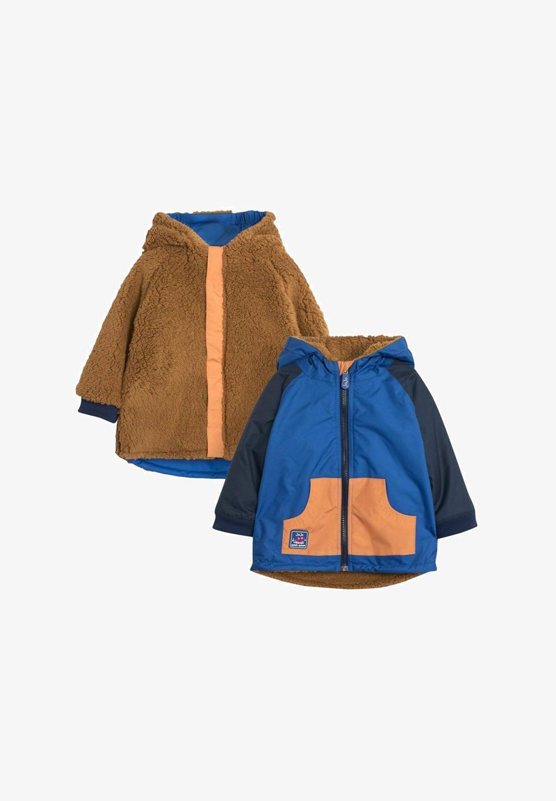 Jachetă din fleece maro cu un fermoar și detalii portocalii, purtată împreună cu o jachetă impermeabilă albastră și bleumarin, având un buzunar portocaliu.