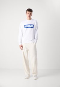 Balta megzti su mėlynu "HUGO" logotipu, su apvalia iškirpte ir gumuotomis rankovėmis. Derinama su laisvomis kreminėmis kelnėmis ir baltomis sportbačiais.