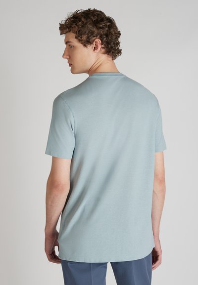 Junger Mann mit lockigem Haar, der ein hellblaues Kurzarmhemd und dunkle Hosen trägt, mit dem Rücken zur Kamera und dem Kopf zur Seite gedreht.