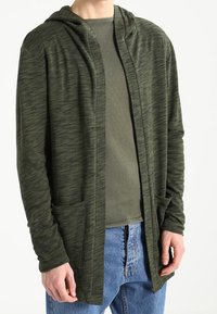 Cardigan lungo aperto in tessuto verde scuro mélange. Presenta un cappuccio, due tasche frontali e polsini a coste. Indossato sopra un top verde chiaro.