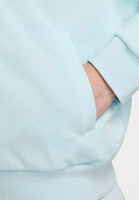 Felpa azzurro chiaro con ampie tasche, polsini a coste e texture morbida. Primo piano che mostra una mano nella tasca.