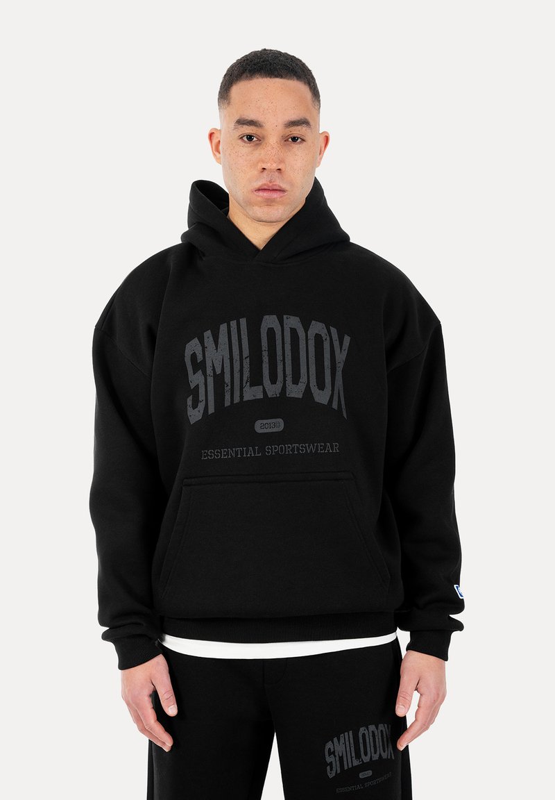 Joven con sudadera con capucha negra y pantalones con el texto "SMILODOX Essential Sportswear", de pie frente a un fondo blanco liso.