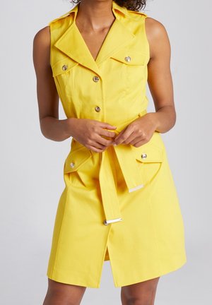 Vrouw in een mouwloze gele jurk met riem, gouden knopen en klepzakken, handen die de riem aanpassen, op een effen lichte achtergrond.