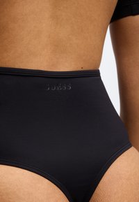 Μαύρο μπικίνι με ψηλή μέση, κατασκευασμένο από λείο ύφασμα, με διακριτικό λογότυπο GUESS σε κομψά γράμματα κατά μήκος της μέσης.