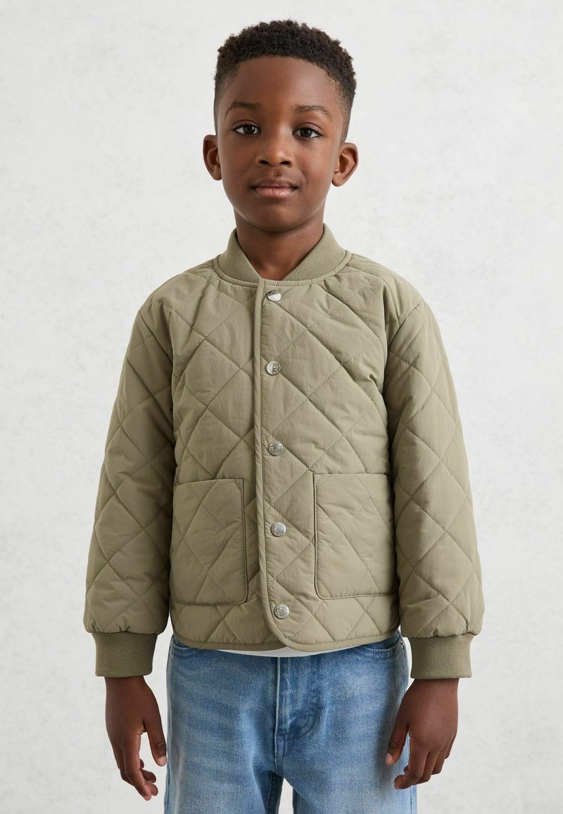 Garçon aux cheveux courts et bouclés portant une veste matelassée vert olive à boutons-pression et un jean bleu clair, debout devant un fond uni et clair.