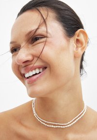 Twee lagen van een delicate witte parelketting, met onregelmatige vormen en een gladde textuur, die naadloos de blote schouders aanvullen tegen een lichte achtergrond.