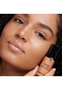 Fluid Sheer glow enhancer van Giorgio Armani, in een glanzend zwarte en koperen fles. Het model heeft een egale huid en natuurlijke make-up, die het product benadrukt.