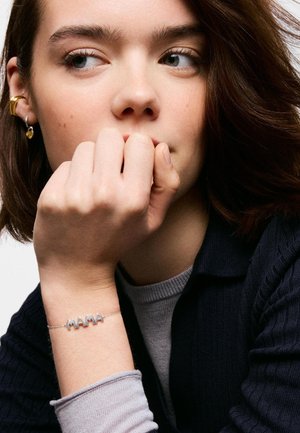 Giovane donna con capelli castani che poggia il mento sulla mano, indossa un braccialetto "MAMA" e orecchini d'oro, guarda di lato con aria pensierosa.