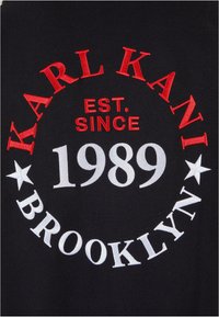 Tejido negro con texto bordado en blanco y rojo. El diseño incluye "KARL KANI", "EST. SINCE 1989" y "BROOKLYN", con acentos de estrellas.