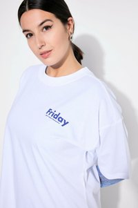 Bílý bavlněný tričko s modrým textem "Pátek" na levé části hrudi. Design má volný střih, krátké rukávy a kulatý výstřih.