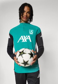 Tamsiai žalsvos ir juodos spalvos sportinis marškinėlis su pusiau užtrauktuku ir balta logotipu, derinamas su juodomis sportinėmis kelnėmis. Laiko baltą futbolo kamuolį su juodais žvaigždžių akcentais.