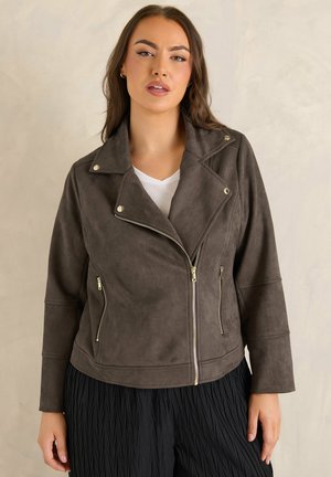 Kvinde iført en mørkegrå ruskind bikerjakke med gyldne lynlåse over en hvid top og sorte teksturerede bukser mod en ensfarvet beige baggrund.