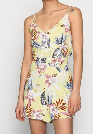 Woman wearing a yellow floral romper with thin straps and a V-neck, featuring pink, blue, white, and brown tropical leaf patterns.