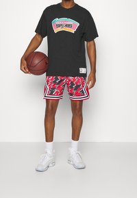Persona che tiene un pallone da basket, indossando una t-shirt nera dei San Antonio Spurs, pantaloni shorts da basket rossi con grafica, calze bianche e scarpe da ginnastica bianche.
