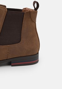 Bottine en daim marron avec panneaux élastiques sur les côtés, petite languette de tirage, et une semelle en caoutchouc contrastante rayée en bleu marine, rouge et noir.