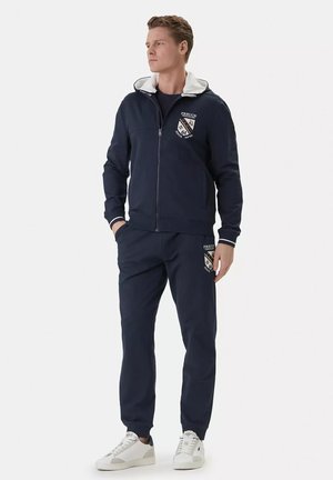 Uomo con felpa con cappuccio blu navy con zip e pantaloni della tuta coordinati con sneakers bianche, in piedi con le mani in tasca, guarda a sinistra.