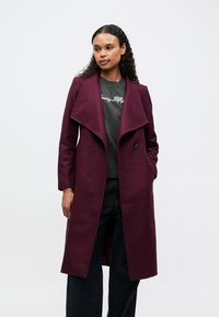 Burgunderfärgad ullblandad kappa med bred krage, enkel knappstängning och sned fåll. Kombinerad med en grå sweatshirt och mörka jeans.