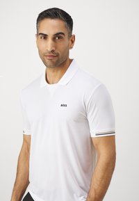 Λευκό πόλο μπλουζάκι με υφασμάτινη υφή, κλασικό γιακά και μικρό μαύρο λογότυπο "BOSS". Ριγέ λεπτομέρειες στους μανσέτες.