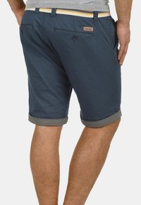 Shorts bleu marine en coton avec des ourlets roulés, une ceinture contrastante beige, des poches arrière et un petit patch logo près de la taille.