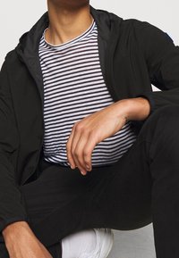 Mann trägt eine schwarze Jacke und Hose, ein schwarz-weiß gestreiftes Hemd, weiße Socken und weiße Turnschuhe, sitzt mit einem angezogenen Knie.