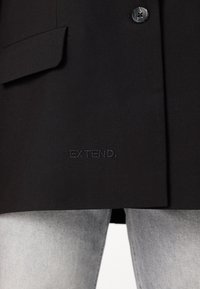 Črn blazer z strukturiranim dizajnom, teksturirano tkanino, zapiranjem na en gumb in subtilno vezenje "EXTEND." na robu.