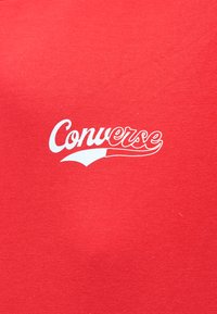 Κόκκινο ύφασμα με το λευκό λογότυπο Converse σε καλλιγραφικά γράμματα, τοποθετημένο στο κέντρο της επιφάνειας.